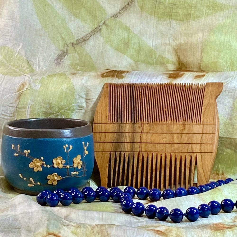 Small Version of Reproduction of Egyptian Comb-Two-Sided, Hand-Carved from Apricot Wood on Silk Background with Blue Beads and Blue Teacup in Foreground