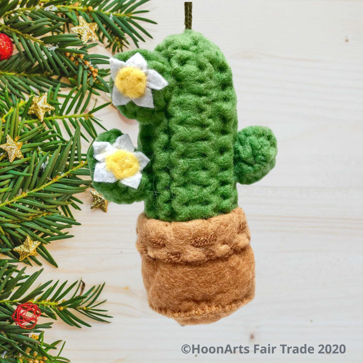 Handmade felt saguara cactus Chrismas ornament, hanging in front of a light background with Christmas branches and tiny decorations on the left of the photo