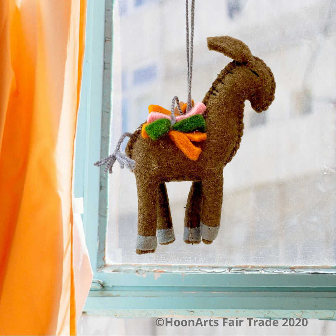 Handmade brown felt donkey ornament from Kyrgyzstan, carrying a load of green, orange and pink felt strips, hanging in front of a window with a blurred view of a tall building, framed by an aqua-colored wooden window frame and a bright apricot-colored curtain to the left | HoonArts