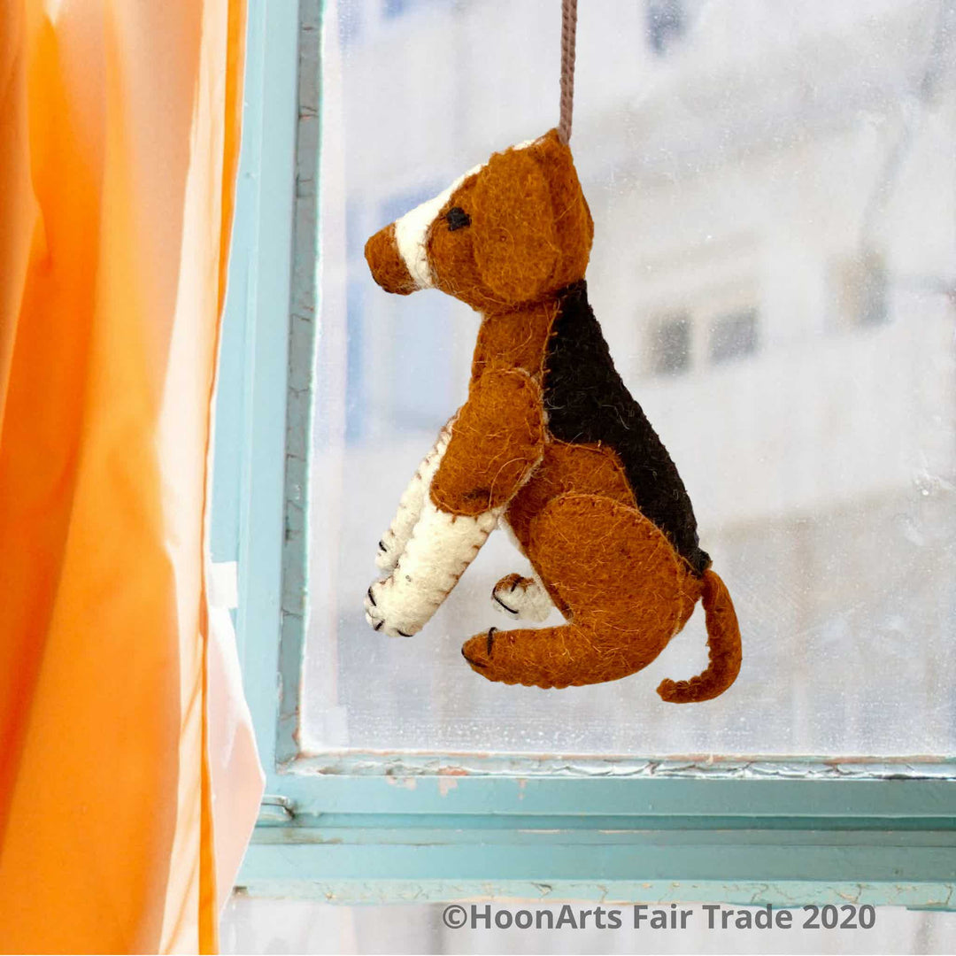 Handmade felt ornament from Kyrgyzstan-Beagle dog with black tan and white patches, seated on hind legs, hanging in front of a window with a blurred view of a multi-story building, framed by an aqua-colored wooden window frame and orange curtain to the left| HoonArts