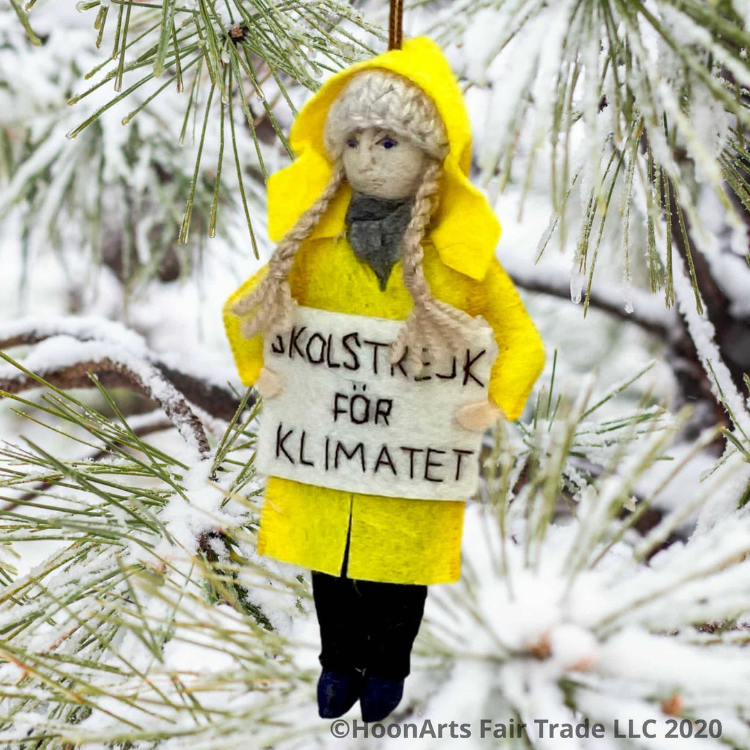 Handmade Felt Ornament of Young Swedish Environmental Activist Greta Thunberg, dressed in bright yellow raincoat and knitted wool hat with long blond brains, carrying a white sign that says "SKOLSTREJK FOR KLIMATET" [school strike for climate]. She is hanging from a snow covered pine tree with long pine needles.