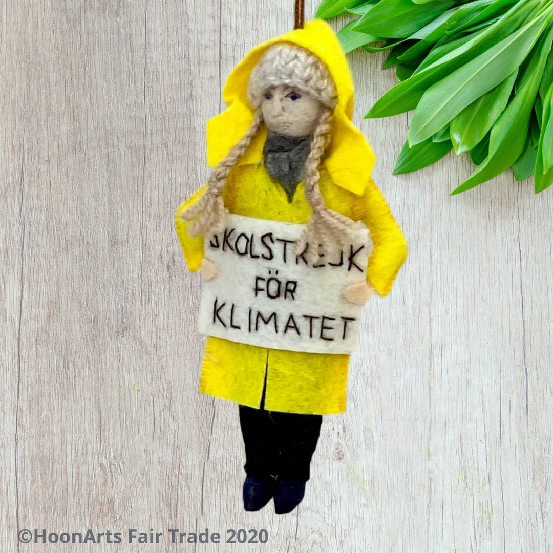 Handmade Felt Ornament of Young Swedish Environmental Activist Greta Thunberg, dressed in bright yellow raincoat and knitted wool hat with long blond brains, carrying a white sign that says "SKOLSTREJK FOR KLIMATET" [school strike for climate]. She is hanging in front of a very light colored wooden wall with a collection of small green leaves  in the upper right corner.