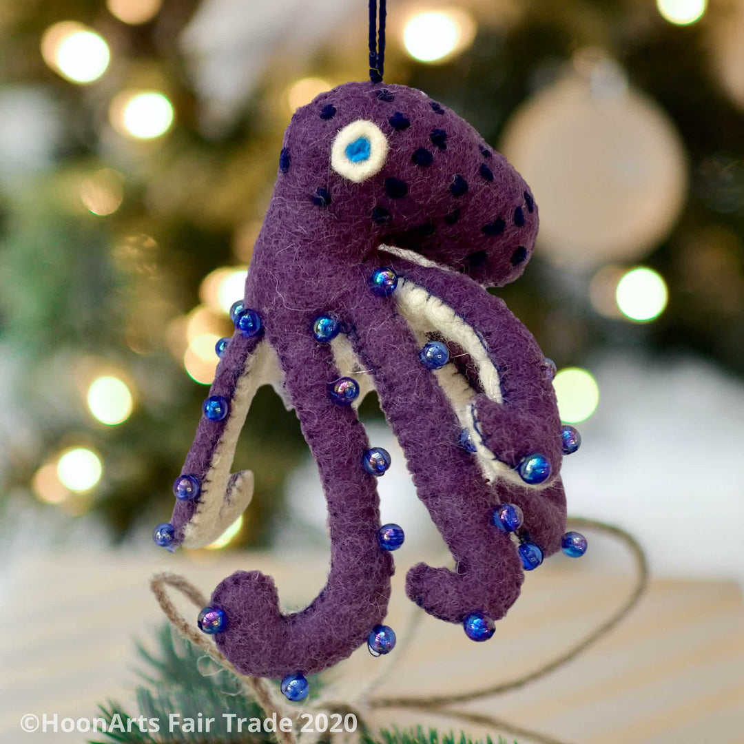 Handmade Felt Ornament-Purple Octopus, with white underside, with blue iridescent beads dotting the purple upper side and bright blue eyes, hanging in front of a blurred image of a Christmas tree with  white lights.