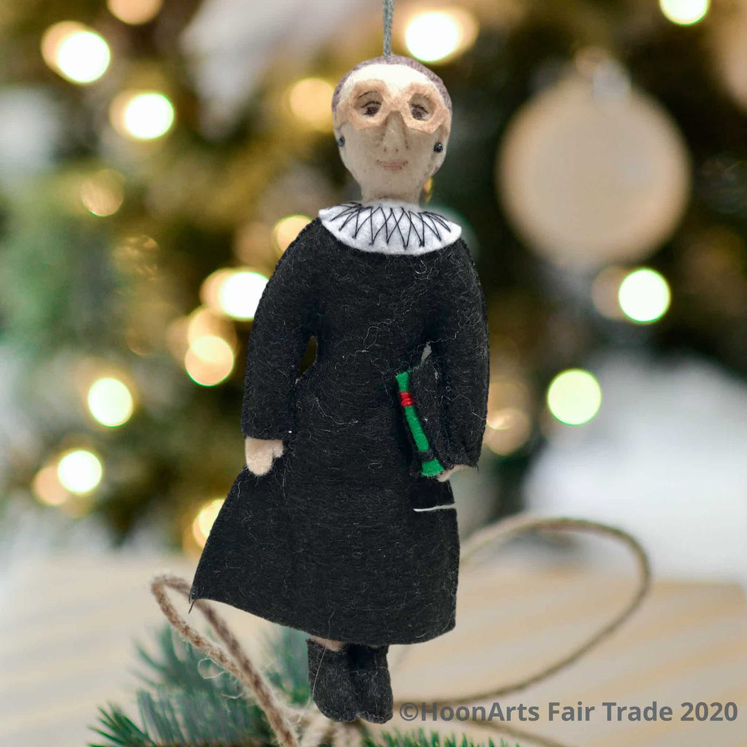 Handmade Felt Ornament-RBG Wearing Black Judicial Robes with Big White Color and Large Eyeglasses, Carrying a Law Book, Hanging against a blurred image of a brightly lit Christmas tree