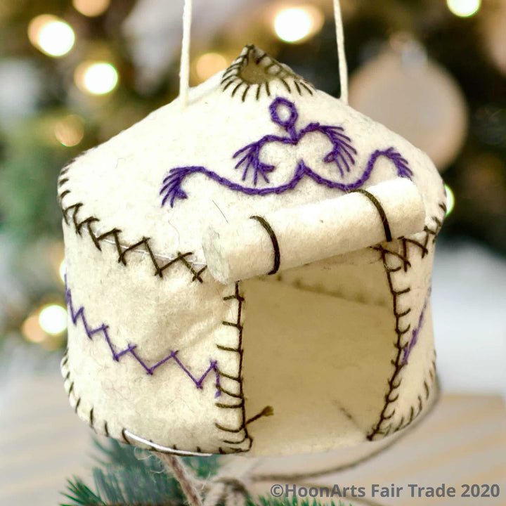 Handmade Felt Yurt Christmas Ornament, White with purple and green hand stitched accents, and doorway rolled up to the roof, hanging in front of a blurred background of a Christmas tree with white lights| HoonArts