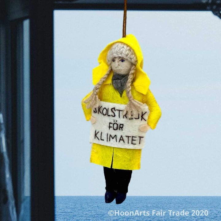 Handmade Felt Ornament of Young Swedish Environmental Activist Greta Thunberg, dressed in bright yellow raincoat and knitted wool hat with long blond brains, carrying a white sign that says "SKOLSTREJK FOR KLIMATET" [school strike for climate]. She is hanging from a blue window frame looking out over a calm blue ocean.