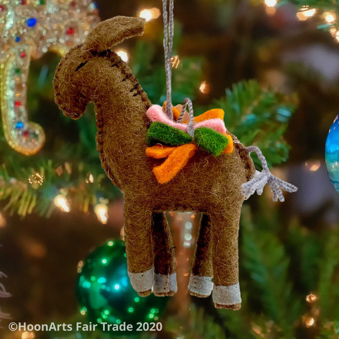Handmade brown felt donkey ornament from Kyrgyzstan, carrying a load of green, orange and pink felt strips, hanging from a Christmas trees with brightly colored ornaments | HoonArts