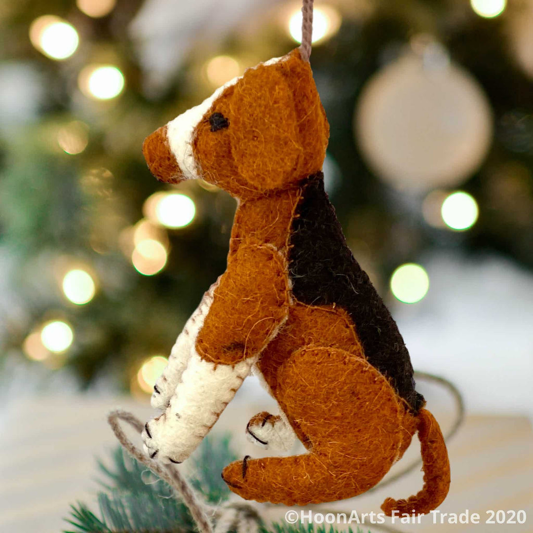 Handmade felt ornament from Kyrgyzstan-Beagle dog with black tan and white patches, seated on hind legs, hanging in front of a blurred image of  a Christmas tree with bright white lights | HoonArts