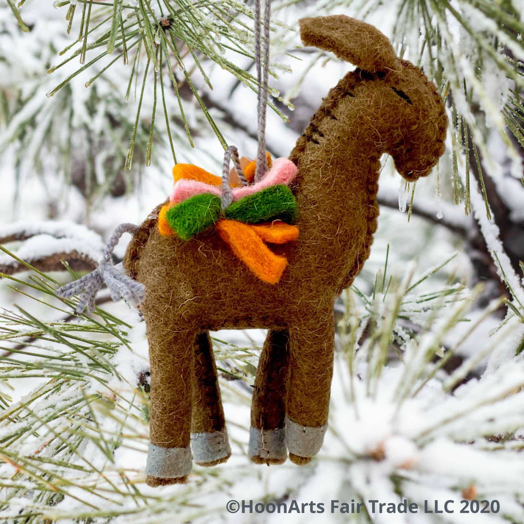 Handmade brown felt donkey ornament, carrying a load of green, orange and pink felt strips, hanging from a snow covered pine tree with long needles | HoonArts