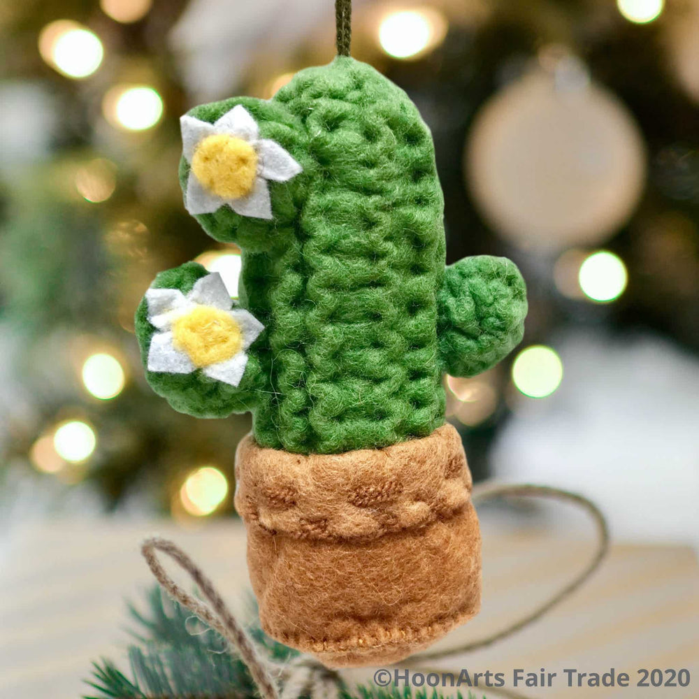 Bright green handmade felt Saguaro cactus Christmas ornament, with two white & yellow blossoms, hanging in front of a blurred background of Christmas twinkle lights