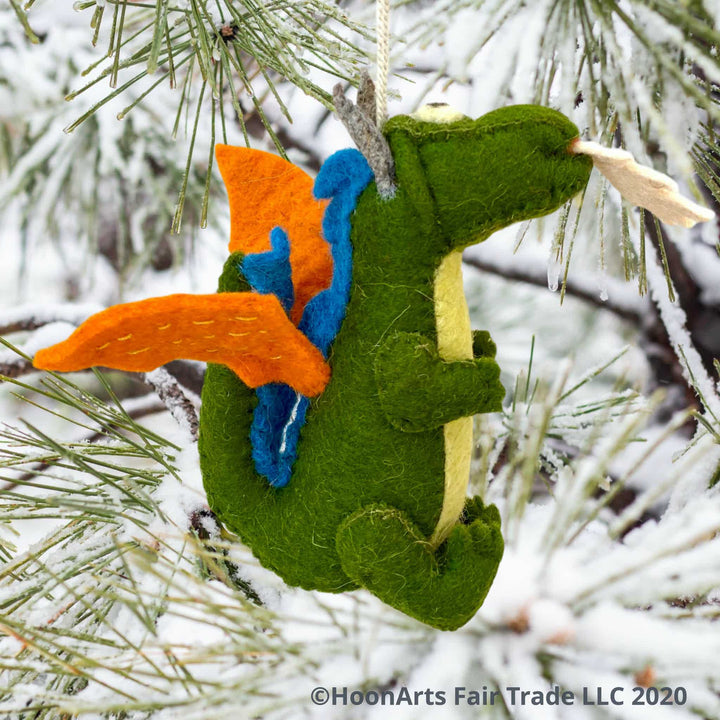 Side view of Handmade Kyrgyz green felt dragon ornament with orange wings, blue accents along the middle of the back,  big black eyes on white and white "flames" shooting from mouth, hanging from a snow covered pine tree with long pine needles | HoonArts  