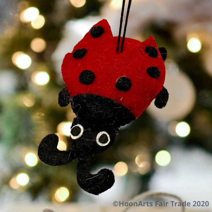 Bright red and black felt ladybug Christmas ornament, hanging in front of a blurred background of Christmas twinkle lights