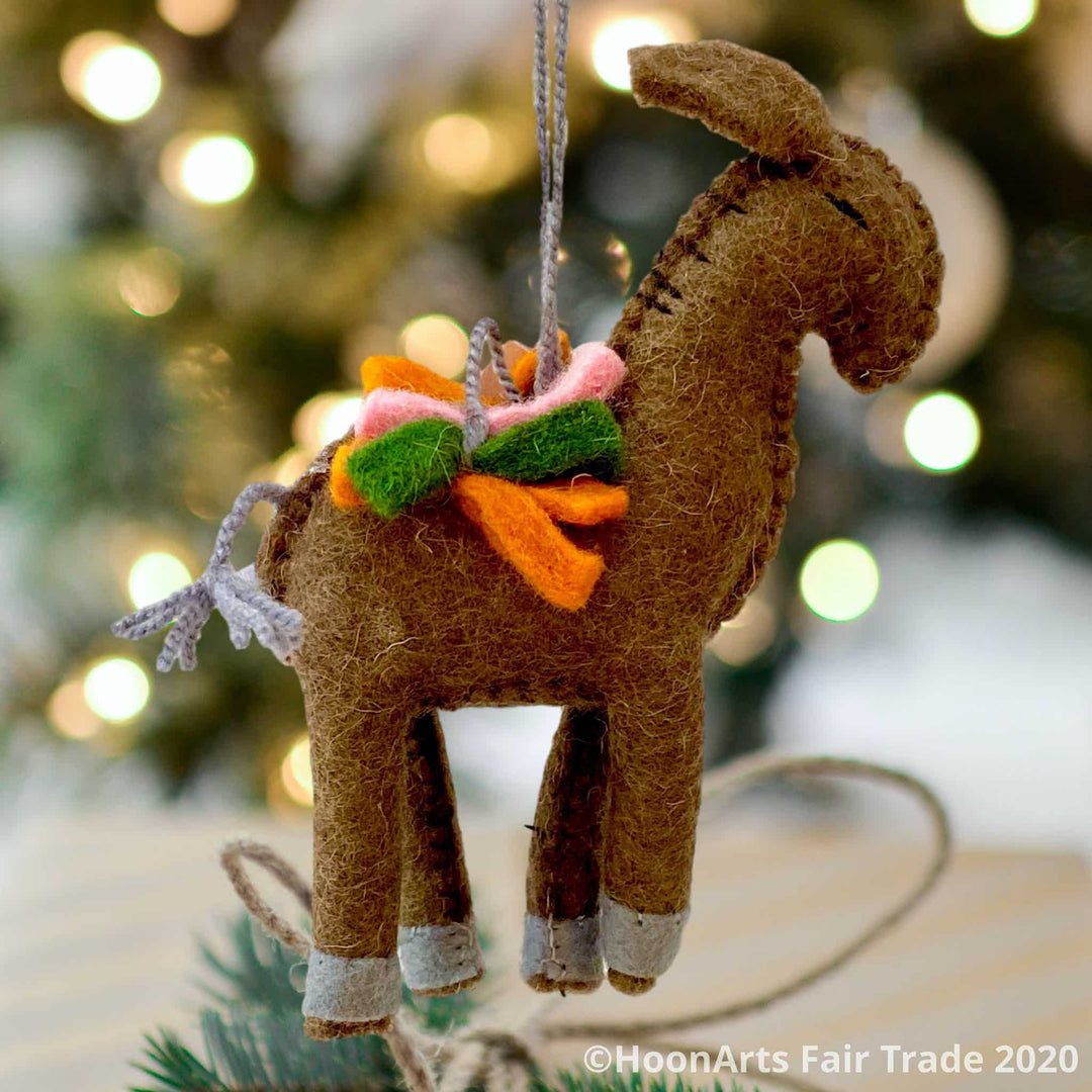 Handmade brown felt donkey ornament from Kyrgyzstan, carrying a load of green, orange and pink felt strips, hanging in front of a blurred image of a Christmas tree with bright white lights | HoonArts
