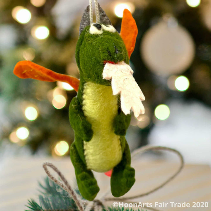 Handmade Kyrgyz green felt dragon ornament with orange wings, big black eyes on white and white "flames" shooting from mouth, hanging in front of a blurred image of a Christmas tree with bright white lights | HoonArts