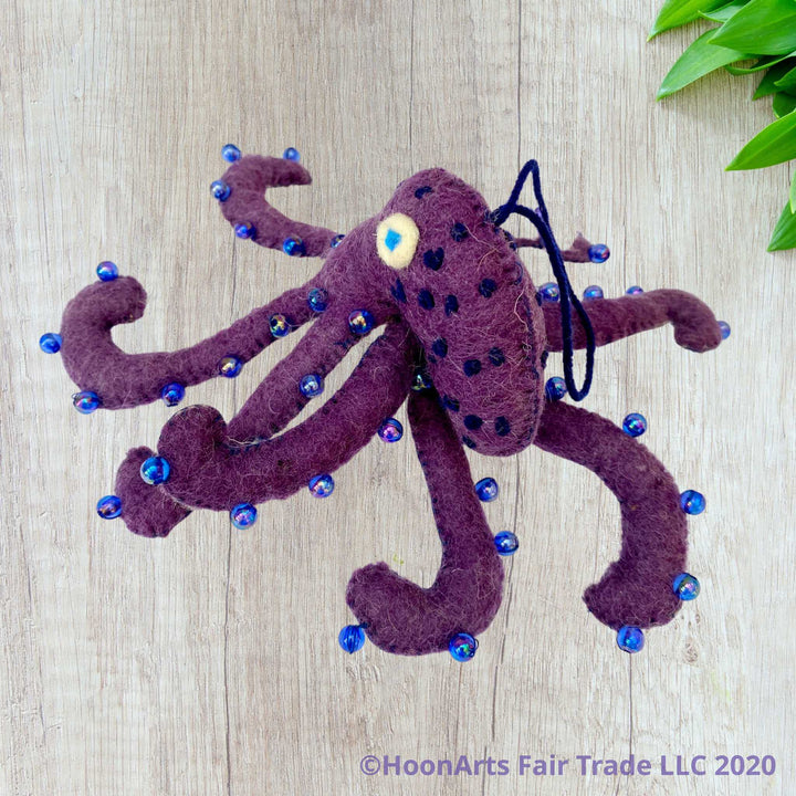Handmade Felt Ornament-Purple Octopus, with white underside, with blue iridescent beads dotting the purple upper side and bright blue eyes, sitting on a white wooden surface with all 8 legs spread out around the head, and small green leaves in the upper right corner of the image.