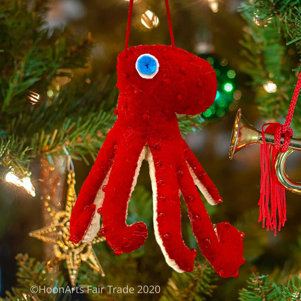 Handmade Felt Ornament-Red Octopus, with white underside, with red beads dotting the red upper side and bright blue eyes, hanging from a Christmas trees with bright ornaments and lights.