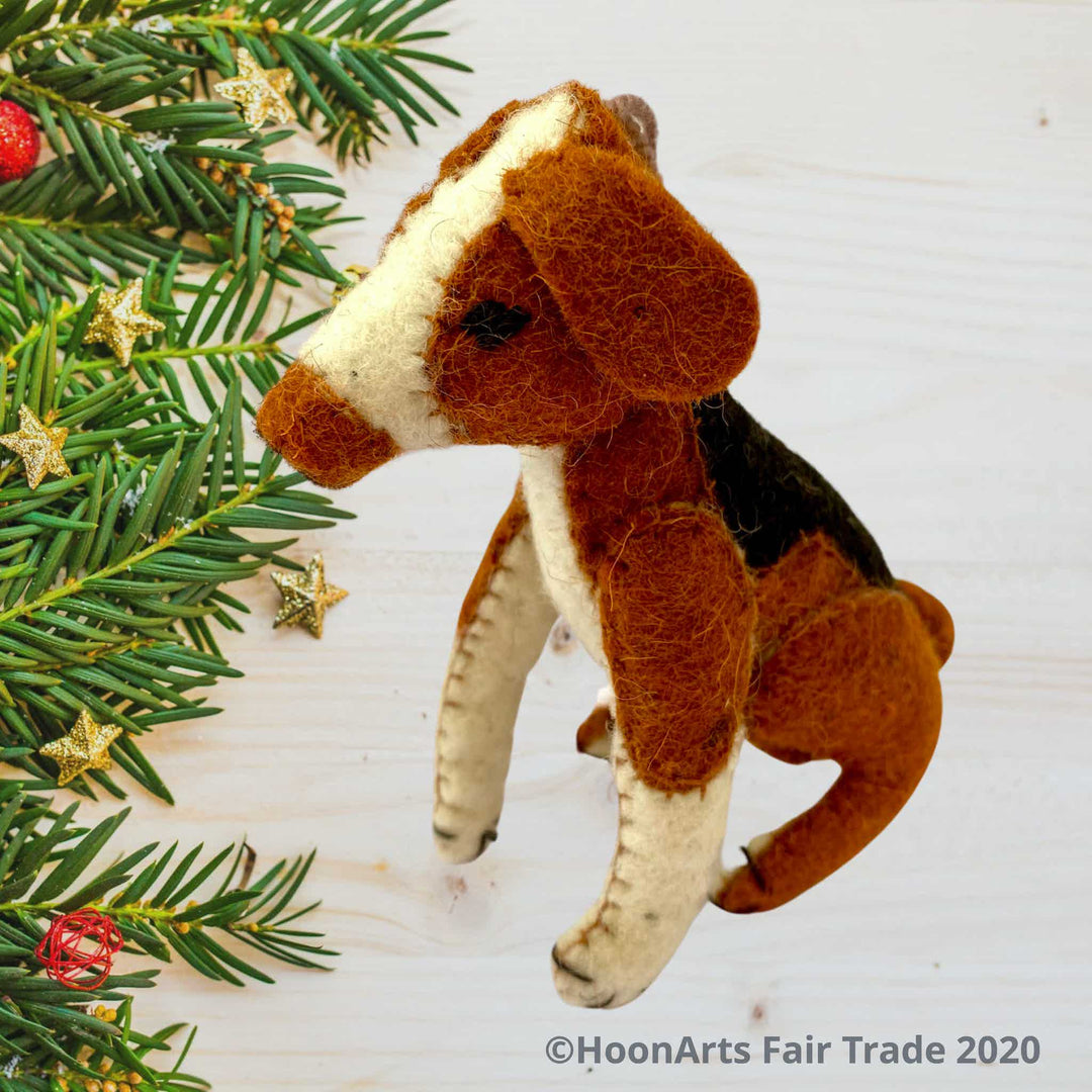 Handmade felt ornament from Kyrgyzstan-Beagle dog with black tan and white patches, seated on hind legs, seated on a whitewashed wooden table with small pine branch tips decorated with small gold starts along the left side of the image| HoonArts