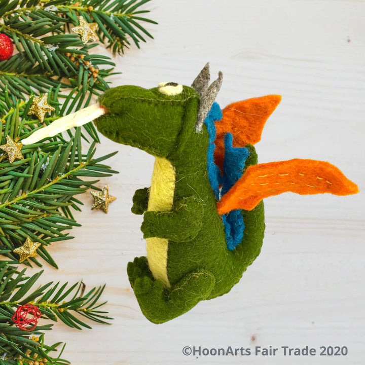 Side view of Handmade Kyrgyz green felt dragon ornament with orange wings, blue accents along the middle of the back, big black eyes on white and white "flames" shooting from mouth, sitting on a white washed wooden table with pine branch tips decorated with tiny gold stars along the left side of the image| HoonArts