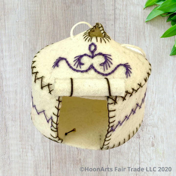 Handmade Felt Yurt Christmas Ornament, White with purple and green hand stitched accents, and doorway rolled up to the roof, sitting on a whitewashed wooden table with bright green leaves inn the upper right corner of the image| HoonArts