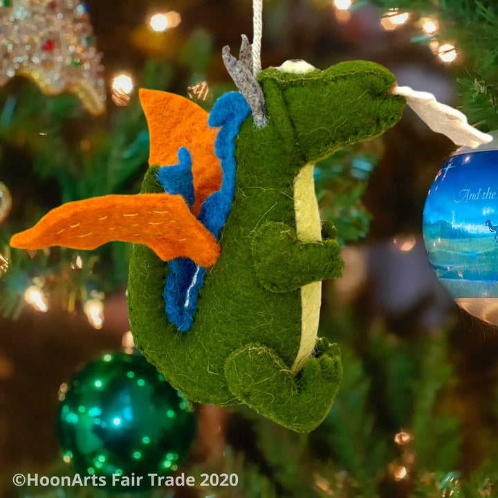 Handmade green felt dragon ornament with orange wings and blue accent along the back, with big eye and white "flames" shooting from mouth, hanging on a Christmas tree with brightly colored bulbs | HoonArts