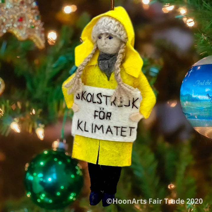 Handmade felt ornament of Greta Thunberg, young environmental activist from Sweden, dressed in Yellow Raincoat, with long blond braids and a knitted wool hat, hanging from a brightly lit Christmas tree. She is carrying a white sign with the words "SKOLSTREJK FOR KLIMATET" [school strike for climate]
