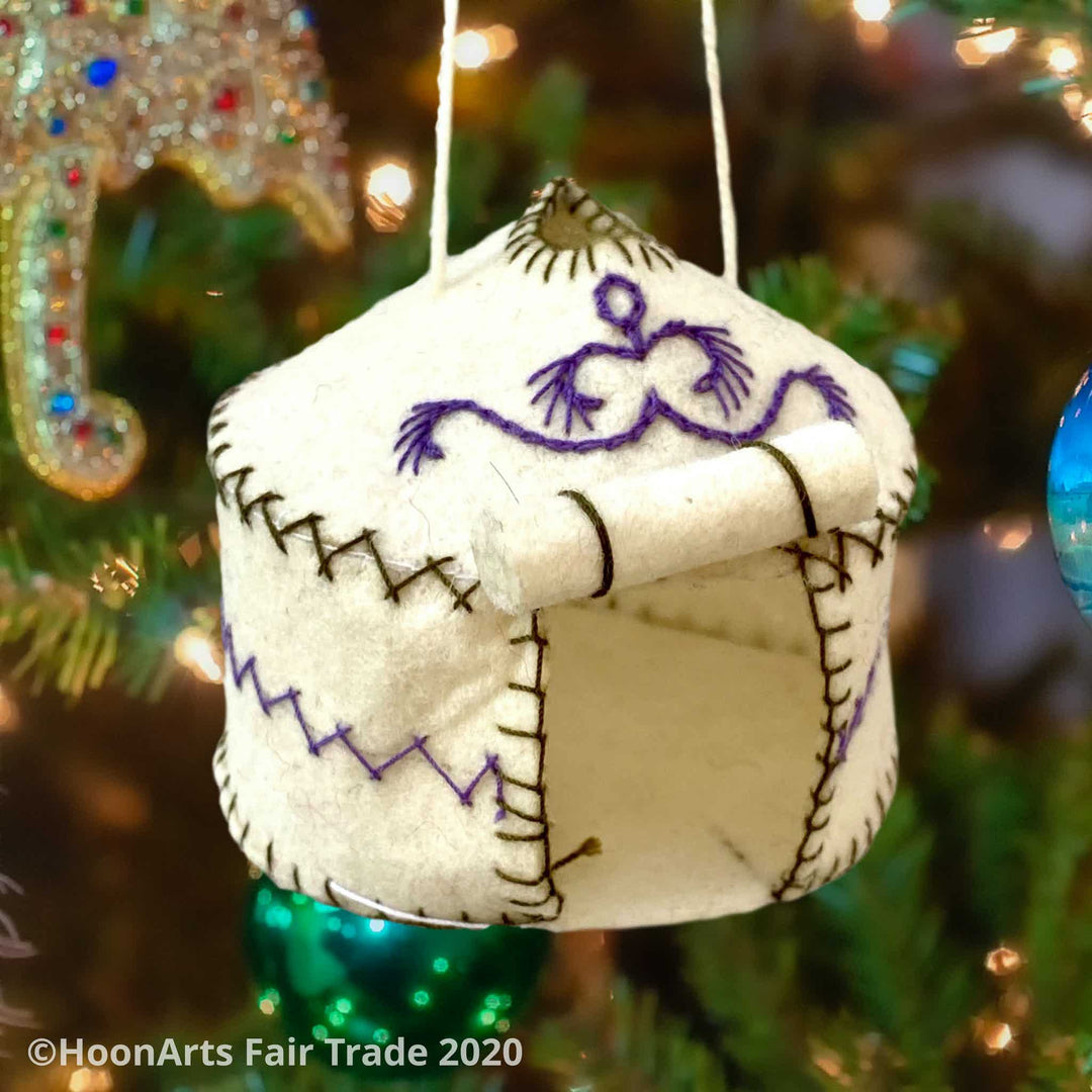 Handmade Felt Yurt Christmas Ornament, White with purple and green hand stitched accents, and doorway rolled up to the roof, hanging from a Christmas tree with brightly colored ornaments and white twinkle lights | HoonArts