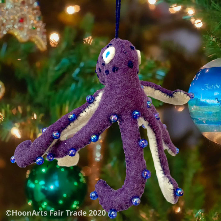 Handmade Felt Ornament-Purple Octopus, with white underside, with blue iridescent beads dotting the purple upper side and bright blue eyes, hanging from a Christmas tree with colorful ornaments and white lights.