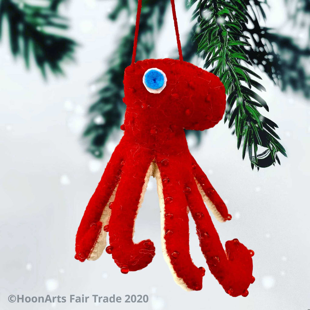 Handmade Felt Ornament-Red Octopus, with white underside, with red beads dotting the red upper side and bright blue eyes, hanging from a pine tree against a white background.