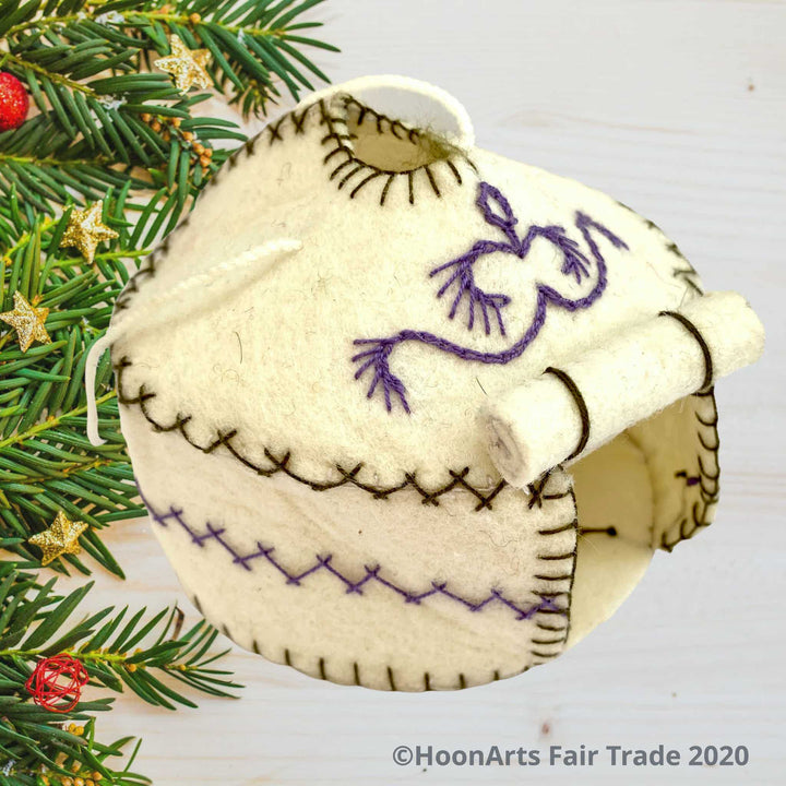 Handmade Felt Yurt Christmas Ornament, White with purple and green hand stitched accents, and doorway rolled up to the roof, sitting on a whitewashed wooden table with pine branch tips decorated with tiny gold stars and red balls along the left side of the image| HoonArts