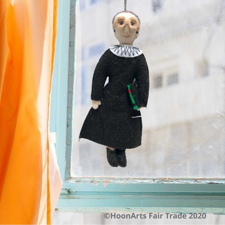 Ruth Bader Ginsburg Handmade Felt Figure, Dressed in Black Judicial Robes and White Color, Wearing Her Signature Big Glasses, Carrying Her Law Book. She's hanging in front of a window with an aqua-colored wooden frame and an apricot colored curtain to the left