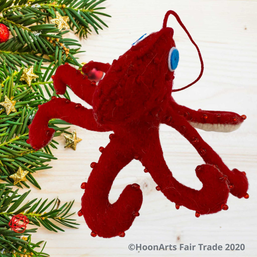 Handmade Felt Ornament-Red Octopus, with white underside, with red beads dotting the red upper side and bright blue eyes, pictured on a very light wooden table with pine tips decorated with small golden stars on the left side of the image.